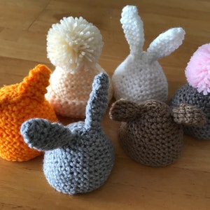 May include: Six crocheted hats in various colors and styles, including a white bunny hat, a pink pom-pom hat, and a gray hat with bunny ears.