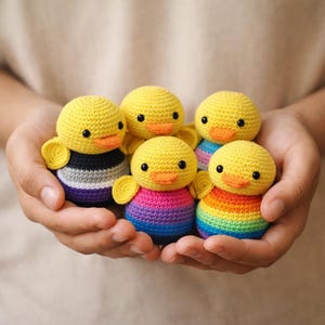 May include: Five small, round, crocheted ducklings with bright yellow heads and orange beaks. Each duckling has a different colored body: one with black, gray, and purple stripes, one with pink and purple, one with rainbow stripes, and two yellow.