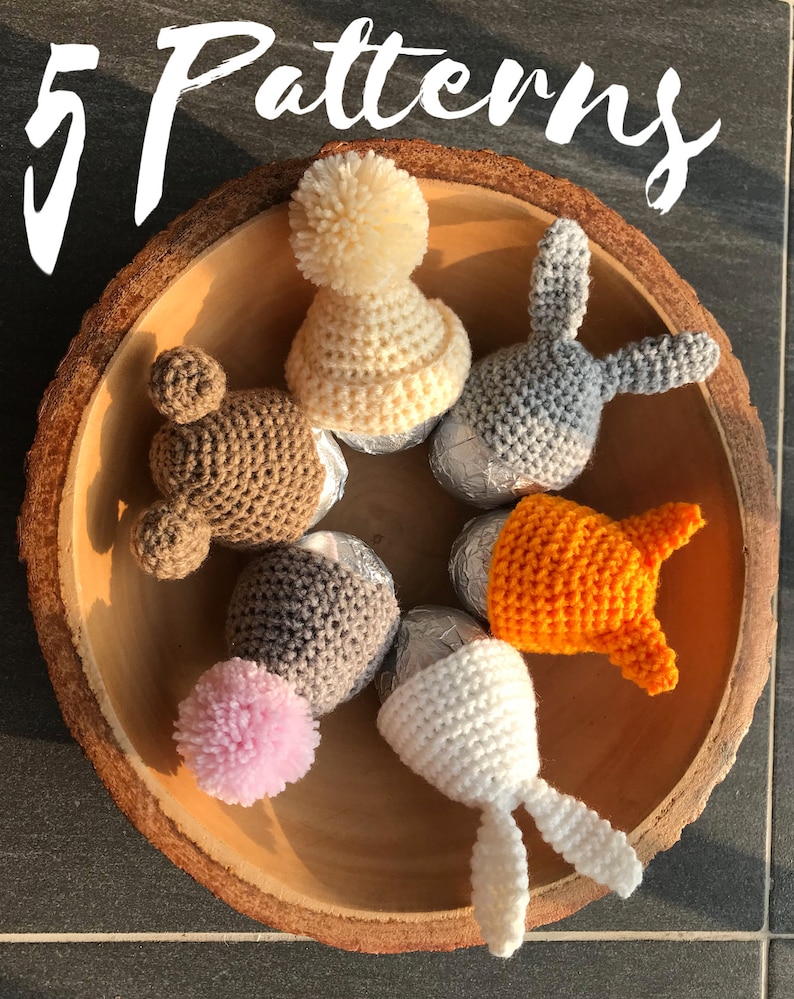 May include: Five crocheted egg cosies in a wooden bowl. The cosies are in the shapes of a bear, bunny, and a hat. The colors are brown, gray, white, orange, and pink.