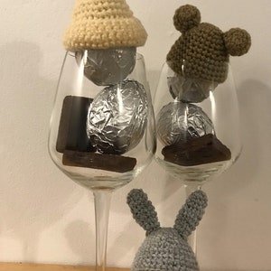 May include: Three chocolate eggs wrapped in foil, each wearing a crocheted hat. The hats are a cream color with a pom-pom, brown with bear ears, and gray with bunny ears.