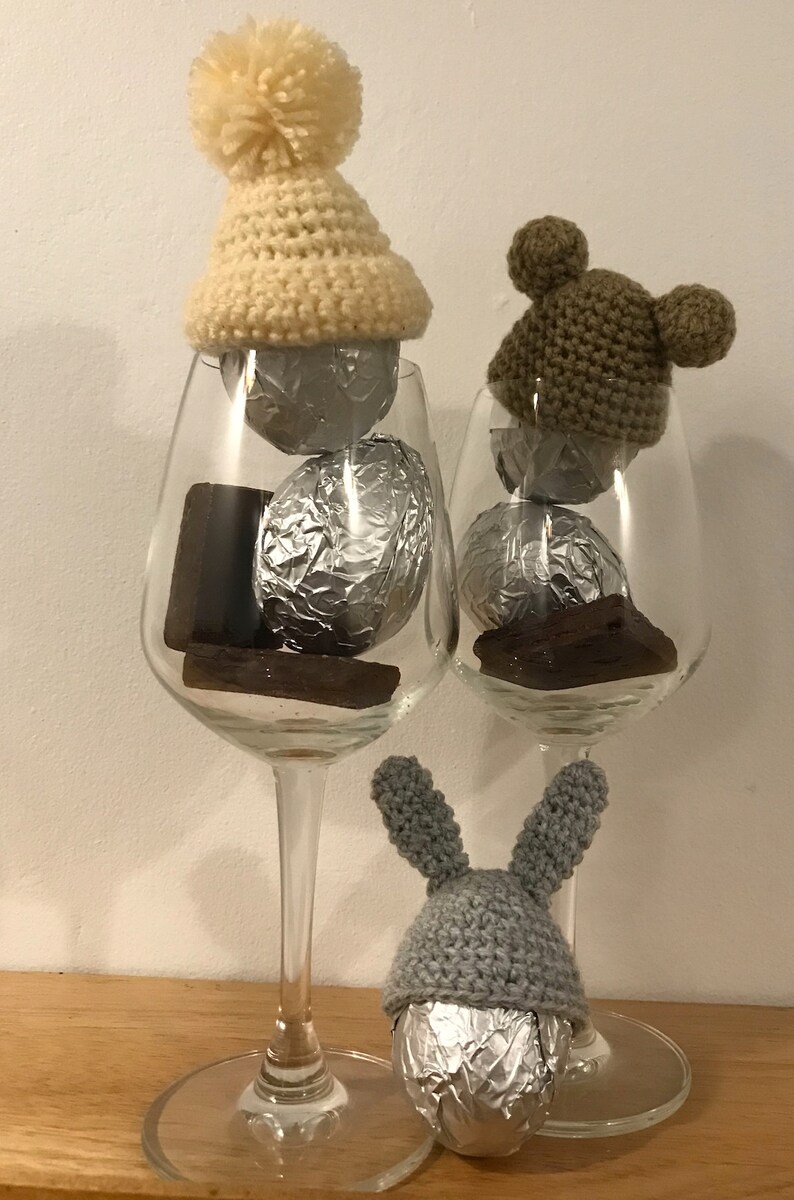 May include: Three chocolate eggs wrapped in foil, each wearing a crocheted hat. The hats are a cream color with a pom-pom, brown with bear ears, and gray with bunny ears.