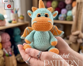 May include: A crocheted dragon toy in shades of teal and orange. The dragon has large black eyes, small wings, and a horn. The image includes the text "crochet PATTERN" and a PDF icon.