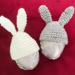 May include: Two crocheted bunny hats in white and gray, each placed on a silver foil-wrapped chocolate egg. The hats are made with a simple stitch pattern and have long, floppy ears.