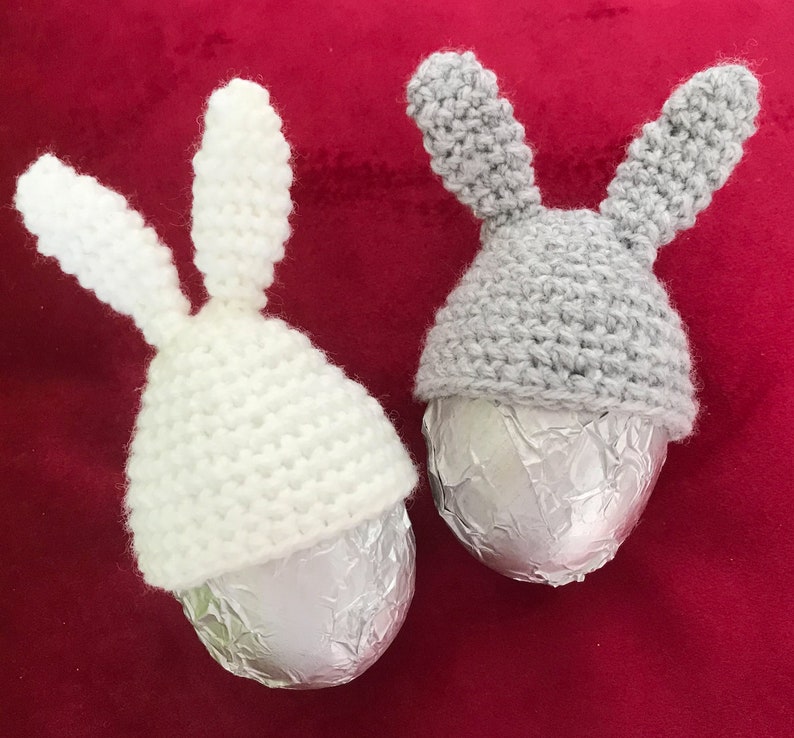 May include: Two crocheted bunny hats in white and gray, each placed on a silver foil-wrapped chocolate egg. The hats are made with a simple stitch pattern and have long, floppy ears.