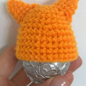 May include: A small orange crocheted hat with two pointy ears, covering a silver foil-wrapped chocolate egg.