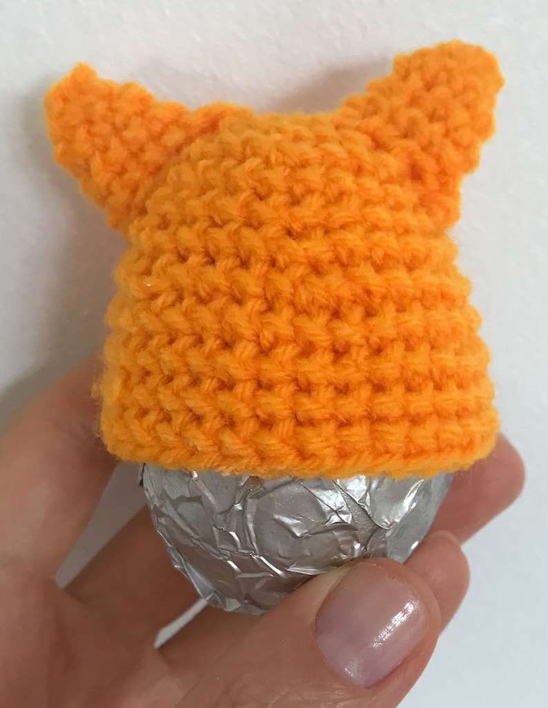 May include: A small orange crocheted hat with two pointy ears, covering a silver foil-wrapped chocolate egg.