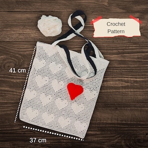 May include: A white crochet tote bag with a heart pattern, measuring 16 inches wide and 14 inches tall. It features a red heart applique and navy blue and white straps. A ball of white yarn and a "Crochet Pattern" sign are also visible.
