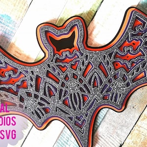 Pode incluir: Um morcego de papel 3D em camadas com um esquema de cores preto, laranja e roxo. O morcego é decorado com um acabamento brilhante e tem um design intrincado.