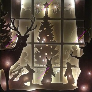 Op de afbeelding: Een houten raamkozijn met een silhouet van een hert, twee konijnen, een kerstboom en een sneeuwpop. Het raam wordt van achteren verlicht met witte lampjes.