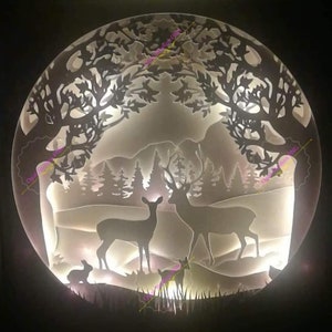 Peut inclure: Une découpe en papier blanche représentant une scène de forêt avec deux cerfs, un lapin et un petit faon. Le motif est éclairé par derrière, créant un effet lumineux.