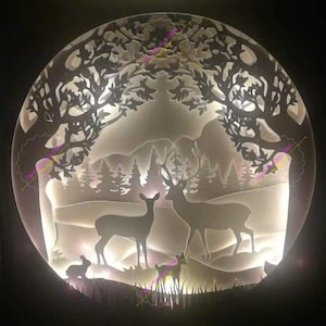 Könnte beinhalten: Ein weißes Papier-Ausschnitt-Design einer Waldszene mit zwei Rehen, einem Kaninchen und einem kleinen Kitz. Das Design wird von hinten beleuchtet, wodurch ein leuchtender Effekt entsteht.