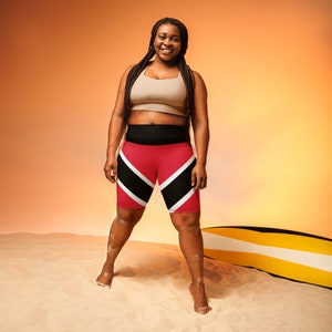 May include: A woman wearing a beige sports bra and red, black, and white athletic shorts with a diagonal pattern. The shorts are designed with the colors of the flag of Trinidad and Tobago. She is standing on a sandy beach with a yellow and black surfboard behind her.