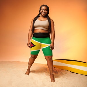 May include: A woman wearing a beige sports bra and black and green athletic shorts with the flag of Guyana printed on them. She is standing on a sandy beach with a surfboard behind her.