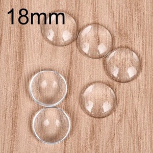May include: Clear, round glass cabochons, each 18mm in diameter, are arranged on a light brown wooden surface. The text "18mm" is displayed in black at the top left. These cabochons are ideal for jewelry making, crafting, or other creative projects.