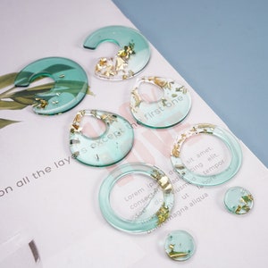Puede incluir: Un conjunto de seis pendientes de resina en varias formas, incluyendo c&iacute;rculos y gotas. Los pendientes son de color verde transl&uacute;cido con escamas doradas incrustadas en la resina. El texto "on all the layers except the first one" y "Lorem ips do sit amet, consectetuadisicing elit, Sect doclusmod tempor ut labore dolore magna aliqua" es visible en el fondo.
