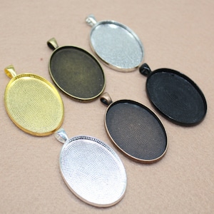 Könnte beinhalten: Fünf ovale Anhänger-Tabletts in verschiedenen Metalloberflächen: Gold, Bronze, Silber, Schwarz und Antik-Silber. Jedes Tablett hat eine strukturierte Oberfläche und eine Öse oben zum Anbringen einer Kette.