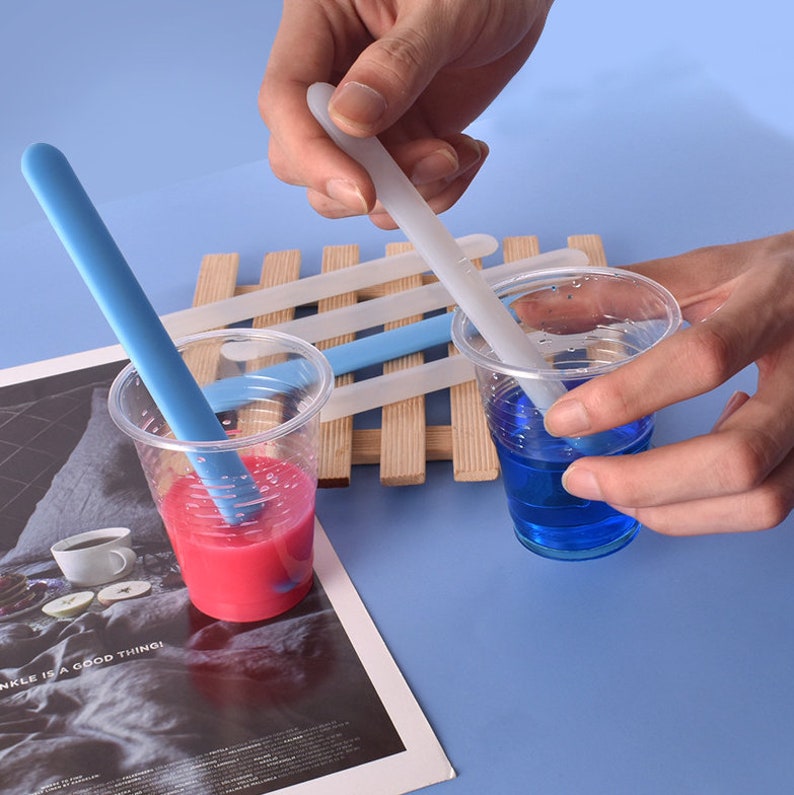 May include: Two clear plastic cups with blue and pink liquid inside. A person is using a white silicone stirring stick to mix the blue liquid. A blue silicone stirring stick is in the pink liquid. There are four white silicone stirring sticks on a wooden surface.