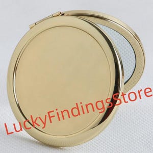 May include: A gold-coloured, round compact mirror, partially open to reveal a reflective surface. The mirror has a shiny, metallic finish. The text "LuckyFindingsstore" is printed in red across the bottom of the mirror.