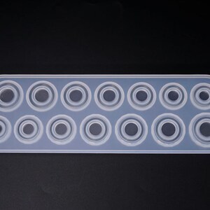Puede incluir: Un molde de silicona transparente de forma rectangular con una serie de cavidades circulares de varios tama&ntilde;os. El molde est&aacute; dise&ntilde;ado para manualidades, probablemente para resina o materiales similares, y tiene una superficie lisa y antiadherente.