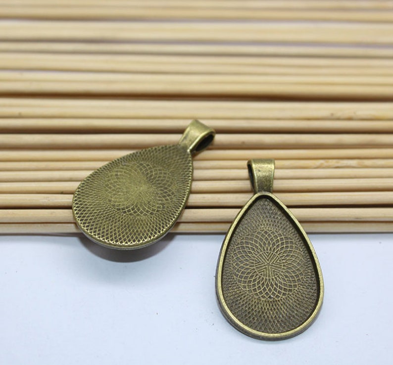 K&ouml;nnte beinhalten: Zwei bronzefarbene Anh&auml;nger-Tabletts in Tropfenform. Die Tabletts haben eine strukturierte Oberfl&auml;che und sind f&uuml;r die Aufnahme eines Cabochons ausgelegt.