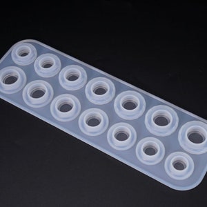 Puede incluir: Un molde de silicona transparente de forma rectangular con m&uacute;ltiples cavidades circulares. El molde est&aacute; dise&ntilde;ado para crear objetos peque&ntilde;os y redondos, con aberturas de diferentes tama&ntilde;os. El fondo es negro liso.