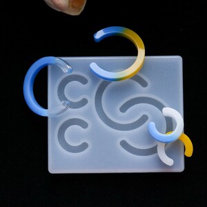 May include: A silicone mold for making resin earrings. The mold has five different shapes, including a crescent moon, a half circle, and a swirl. There are also three resin earrings in the image, one blue and yellow, one blue, and one white.