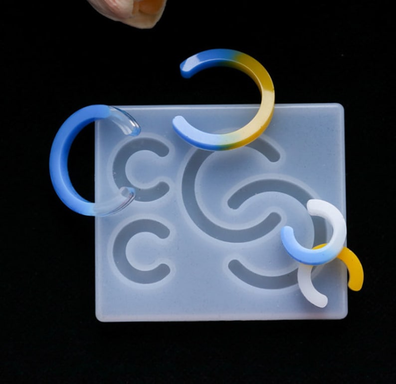 May include: A silicone mold for making resin earrings. The mold has five different shapes, including a crescent moon, a half circle, and a swirl. There are also three resin earrings in the image, one blue and yellow, one blue, and one white.