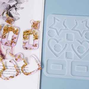 May include: A clear silicone mold for making earrings. The mold has eight different shapes, including stars, hearts, and rectangles. The mold is shown with finished earrings made with resin and gold flakes.