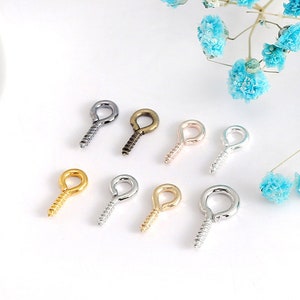 Może przedstawiać: Zestaw ośmiu śrubowych oczek w różnych wykończeniach, w tym srebrnym, złotym, czarnym i brązowym. Oczka są małe i mają zaokrąglony kształt.