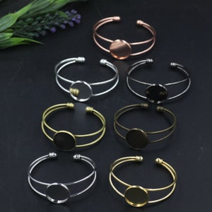 May include: An assortment of open cuff bracelets in various metallic finishes, including silver, gold, bronze, and copper. Each bracelet features a circular setting in the center, perfect for adding a personalized touch. The bracelets are displayed on a dark surface.