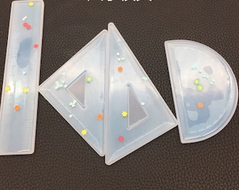 May include: A set of clear resin geometry tools, including a ruler, two triangles, and a protractor. Each tool is embedded with colorful star, flower, and butterfly-shaped glitter. The tools are designed for crafting or educational purposes.