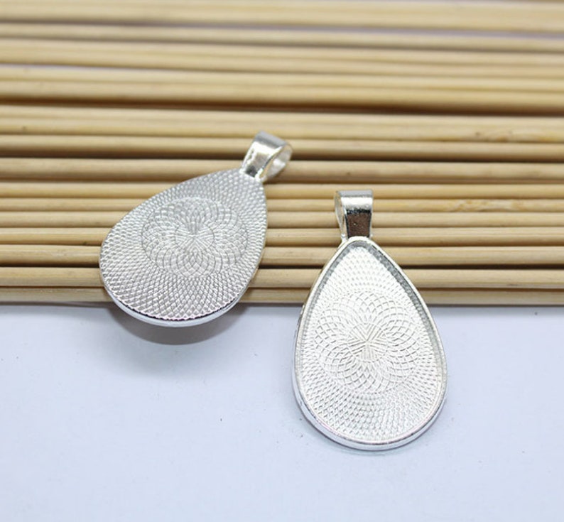 K&ouml;nnte beinhalten: Zwei silberne Anh&auml;nger-Tabletts in Tropfenform mit einer strukturierten Oberfl&auml;che. Die Tabletts sind so konzipiert, dass sie einen flach geformten Cabochon oder ein anderes dekoratives Element aufnehmen k&ouml;nnen.