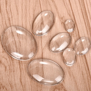 May include: A set of six clear glass oval-shaped cabochons of varying sizes, arranged on a wood surface.