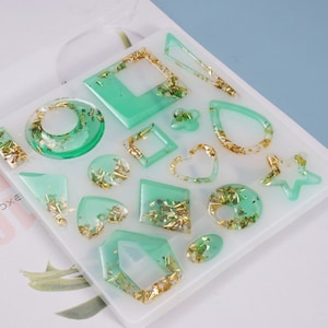 May include: A white silicone mold with 14 different shapes for making resin earrings. The shapes include hearts, stars, circles, squares, and more. The mold is filled with green resin and gold glitter.