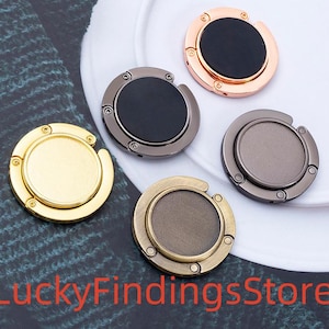 Könnte beinhalten: Fünf metallische Handtaschenaufhänger in Gold, Bronze, Gunmetal, Kupfer und Schwarz. Jeder Aufhänger hat eine runde Oberseite und einen Klapphaken zum Aufhängen von Handtaschen.