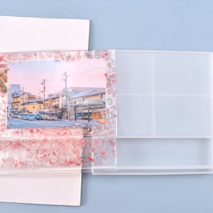 Pode incluir: Uma moldura de resina transparente com uma foto de uma cena de rua. A moldura é decorada com detalhes em rosa e vermelho. Um organizador de resina transparente com vários compartimentos está ao lado da moldura.