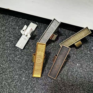 May include: Four different metal rings with a rectangular design. The rings are silver, gold, bronze, and black. Each ring has a textured surface.