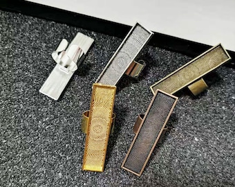 May include: Four different metal rings with a rectangular design. The rings are silver, gold, bronze, and black. Each ring has a textured surface.