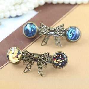 May include: Two gold-toned hair clips with decorative bows. One clip features a blue glass cabochon with a zodiac symbol, the other features a black glass cabochon with a pattern of gold hearts.