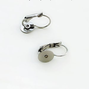 Pode incluir: Duas costas de brincos prateadas são exibidas contra um fundo branco. Cada costas tem uma base circular com um pequeno orifício, uma alavanca articulada e um laço de arame curvo para prender brincos. O metal tem uma superfície brilhante e reflexiva.