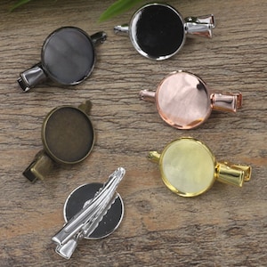May include: Assortment of six silver, gold, and bronze hair clips with round, blank, metal bases. The bases are approximately 2.5 cm in diameter.