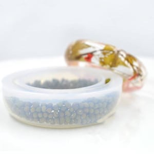 May include: A clear silicone mold for making resin jewelry. The mold is shaped like a ring and has a textured surface. The mold is filled with small, dark brown beads.