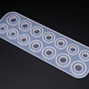 Puede incluir: Un molde de silicona transparente y rectangular con m&uacute;ltiples cavidades circulares de varios tama&ntilde;os. El molde est&aacute; dise&ntilde;ado para crear resina u otros materiales en objetos con forma de anillo. Las cavidades est&aacute;n dispuestas en filas.