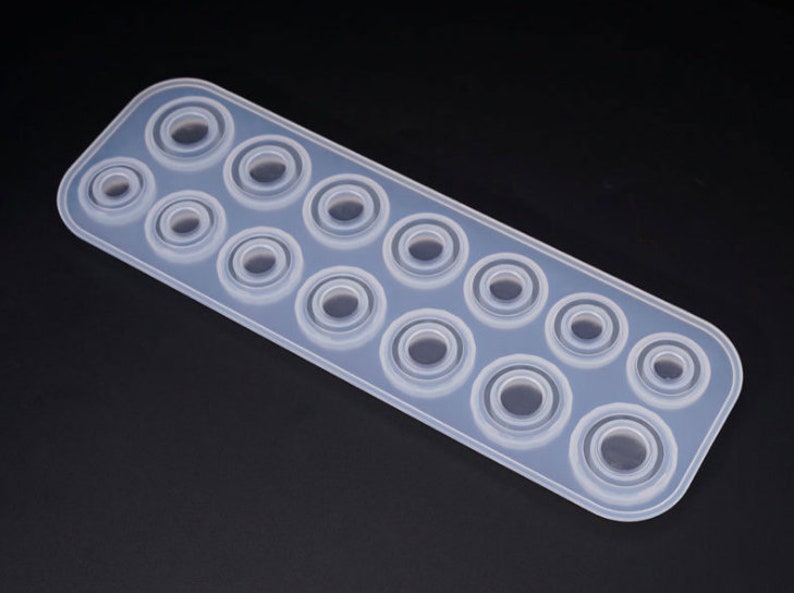 Puede incluir: Un molde de silicona transparente y rectangular con m&uacute;ltiples cavidades circulares de varios tama&ntilde;os. El molde est&aacute; dise&ntilde;ado para crear resina u otros materiales en objetos con forma de anillo. Las cavidades est&aacute;n dispuestas en filas.