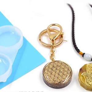 May include: Two resin pendants, one with a gold flower of life design and the other with a gold hamsa hand design. The pendants are attached to a keychain and a necklace with brown cord and white beads.