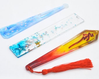 Pode incluir: Três marcadores de livros decorativos com designs diferentes. O primeiro marcador é azul claro com um padrão de floco de neve branco. O segundo marcador é transparente com glitter azul, branco e dourado e uma estrela do mar. O terceiro marcador é vermelho e amarelo com uma borla dourada e um caractere chinês.