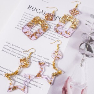 May include: Three pairs of earrings with gold leaf accents encased in clear resin. The earrings are shaped like a rectangle, a circle, and a star. The rectangle and star earrings are pink, while the circle earring is a light purple.
