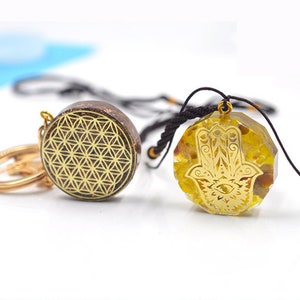 May include: Two gold-toned pendants on black cords. One pendant features a geometric pattern, the other features a hamsa hand with an eye in the center. Both pendants are set in clear resin.