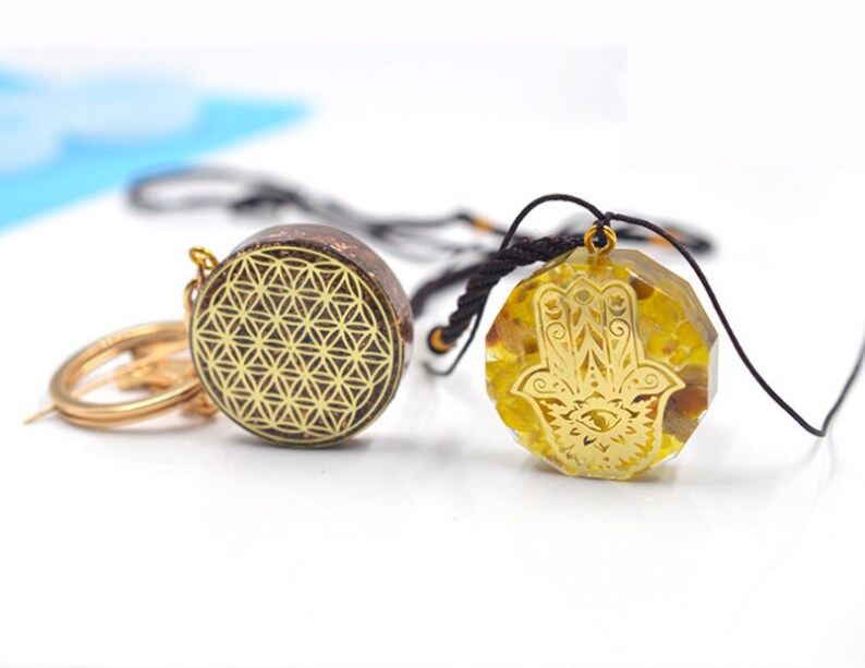 May include: Two gold-toned pendants on black cords. One pendant features a geometric pattern, the other features a hamsa hand with an eye in the center. Both pendants are set in clear resin.