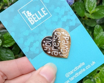 May include: Gold heart-shaped enamel pin with the text "You are so loved" in white lettering. The pin is on a blue background with white hearts.